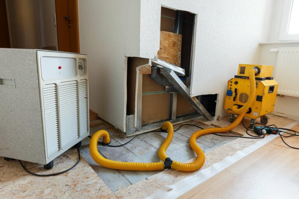 Dehumidifier and air mover connected by yellow hoses in a room under renovation.