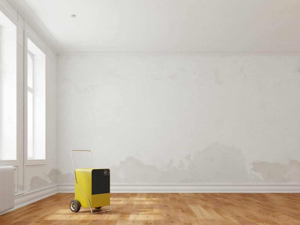 Yellow portable dehumidifier on wheels in an empty room with peeling white walls and wooden floor.
