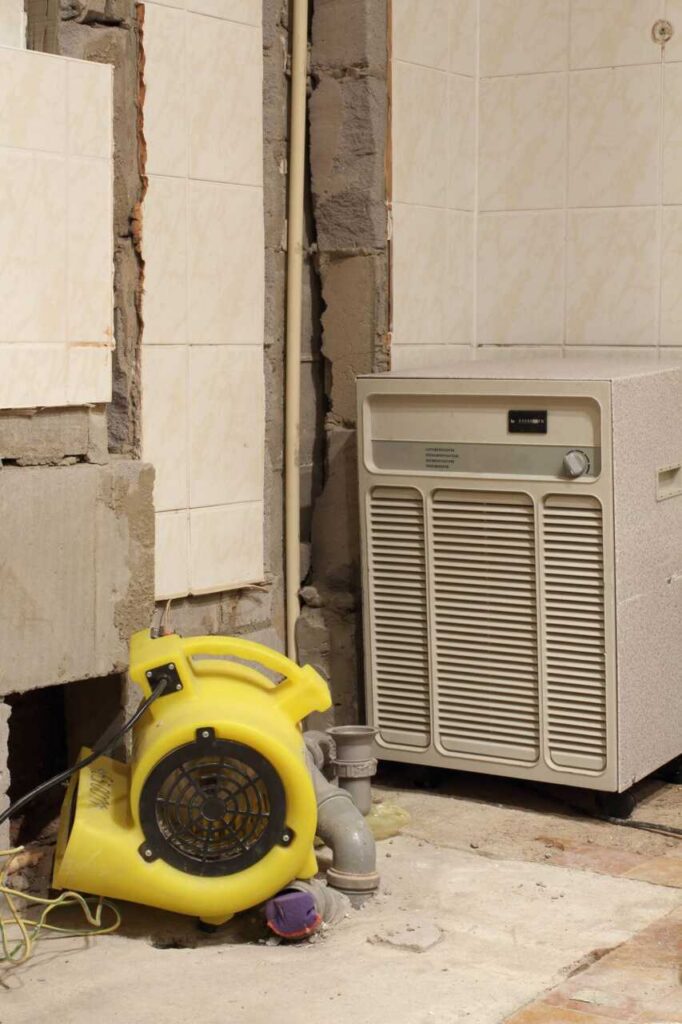 Yellow air blower and beige air conditioning unit against a tiled wall with exposed pipes.