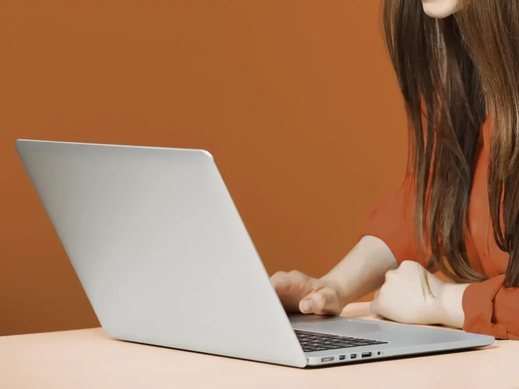 Person using a silver laptop on a light-colored table with an orange background.