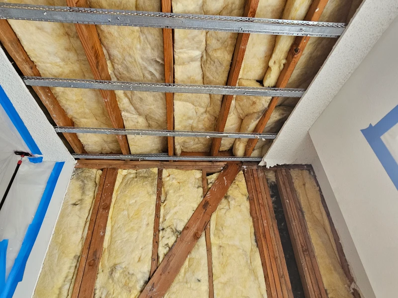 Exposed ceiling and wall insulation with wooden beams and metal framing before storm damage restoration.